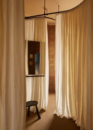 Interior retail fitting room with white fabric curtains, a wall-mounted mirror, and a dark small stool on a carpeted floor.