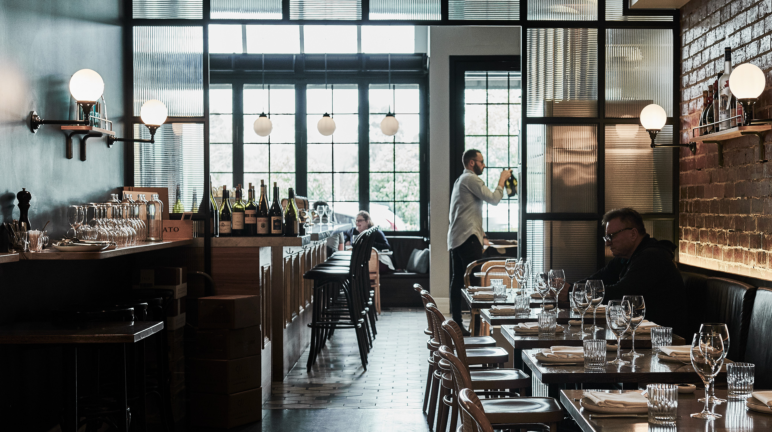 Interior view of a restaurant with tables set, wine bottles on display, and staff attending to patrons in a warmly lit space.