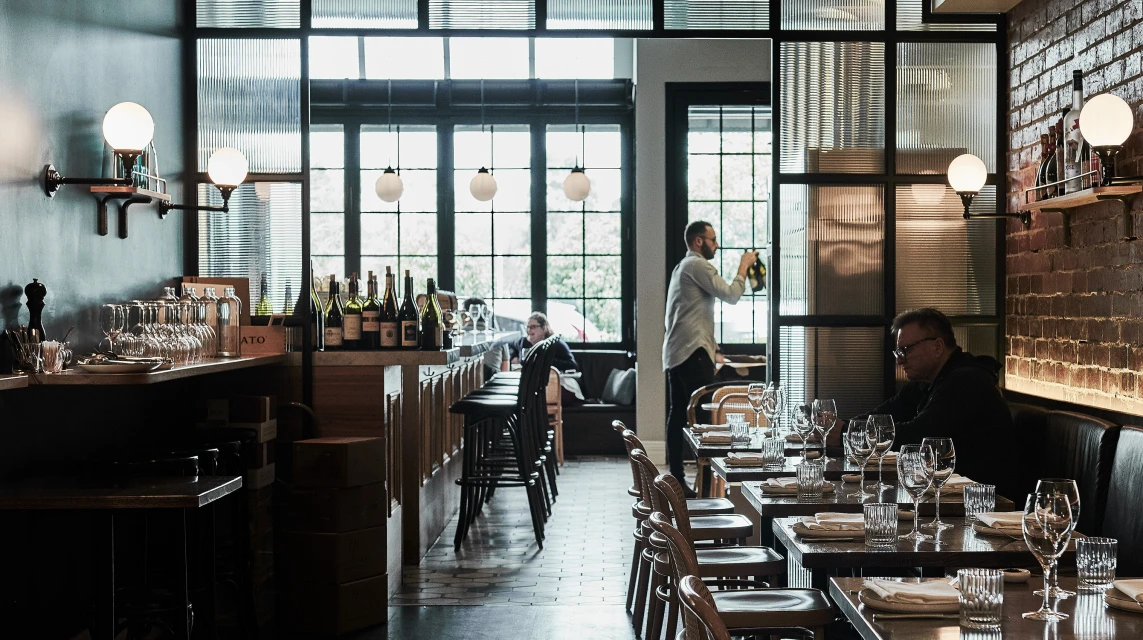 Interior view of a restaurant with tables set, wine bottles on display, and staff attending to patrons in a warmly lit space.