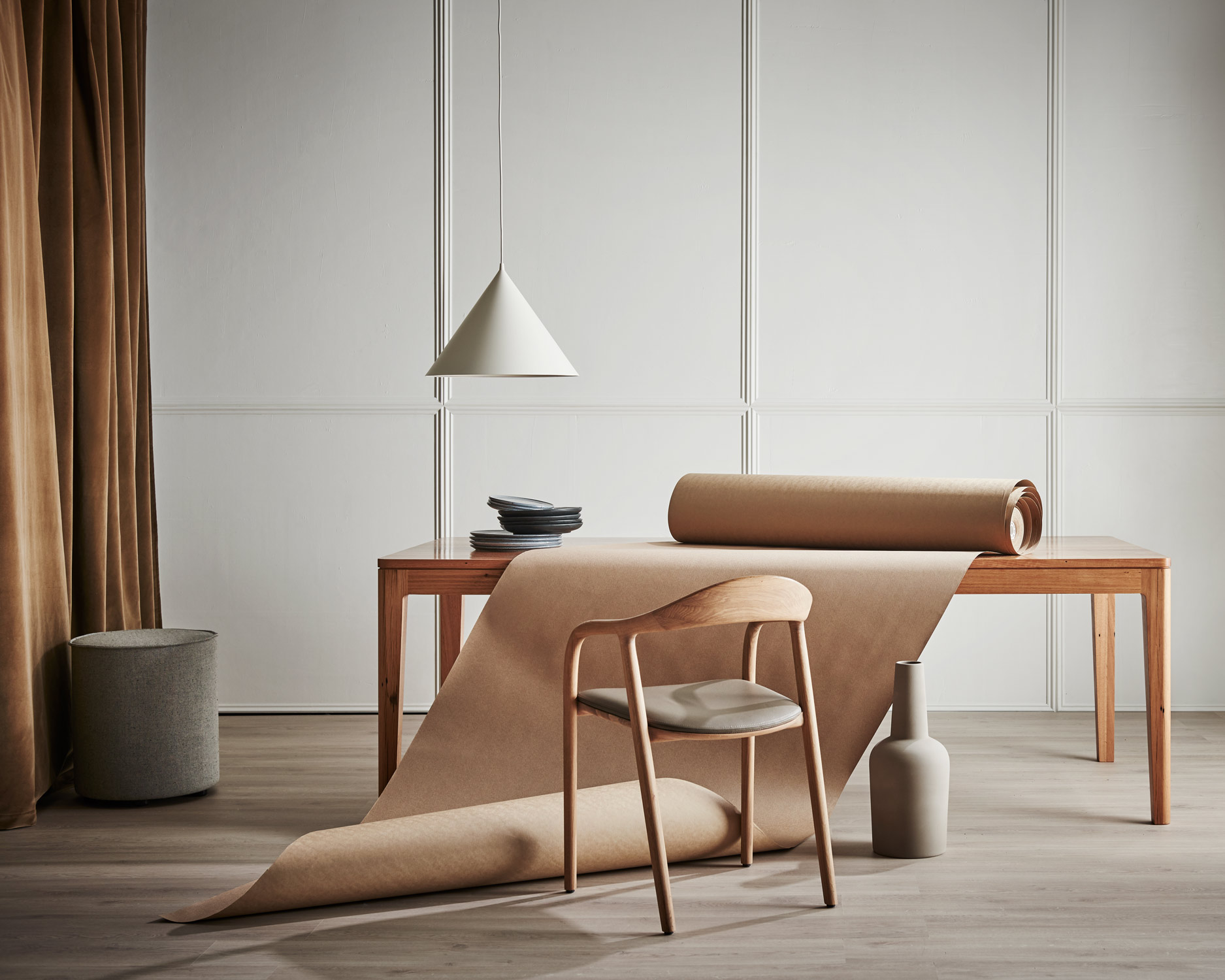 Minimalist interior featuring a wooden chair, table, pendant lamp, and decorative objects against a neutral backdrop with warm brown accents.