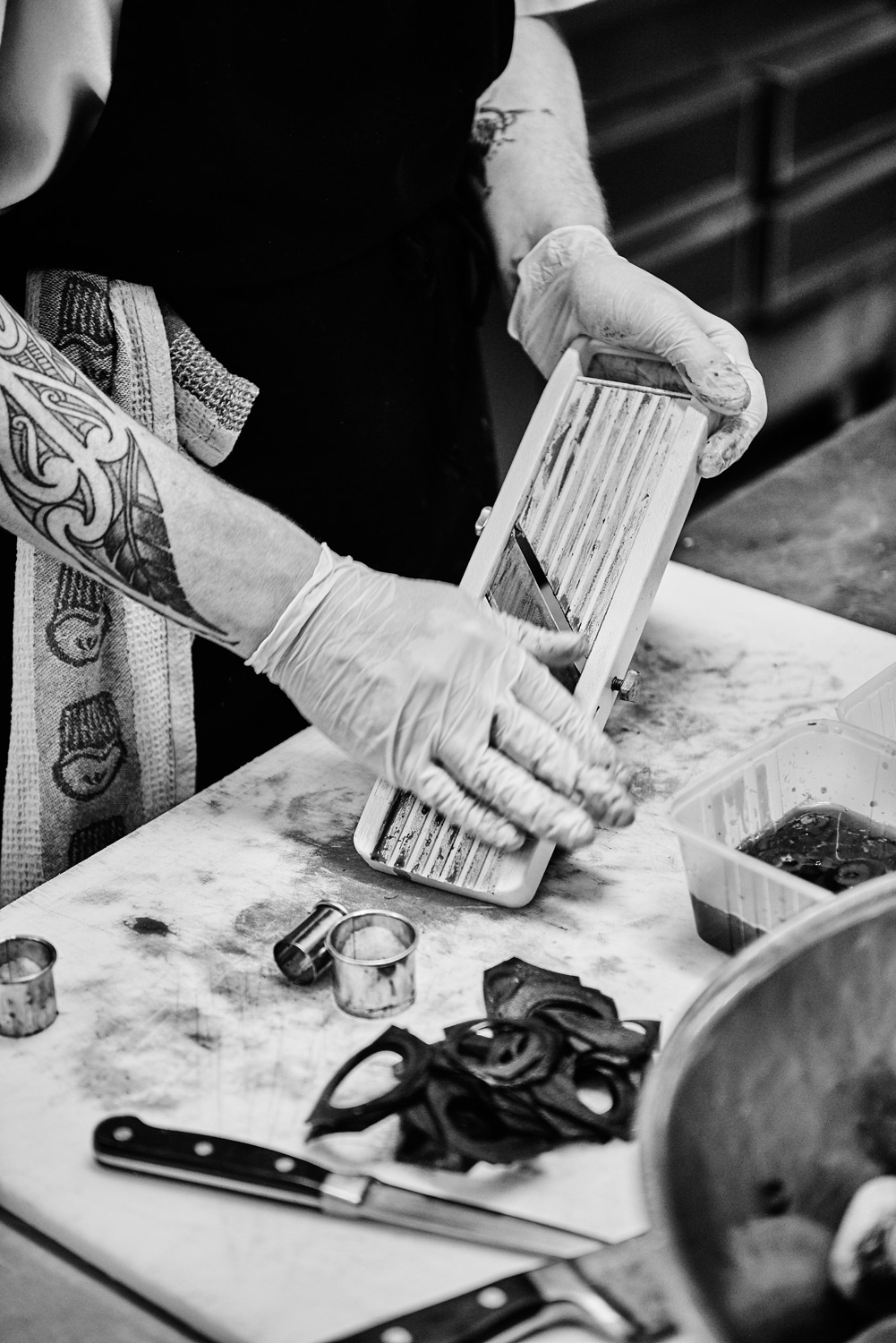 Close-up black and white shot of a chef with tattoos wearing gloves using a mandoline to slice a vegetable with sliced vegetables nearby.