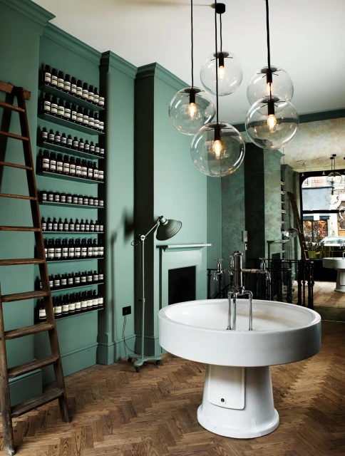 Interior of a store featuring a round sink, globe pendant lights, and shelves stocked with bottles against a teal wall.