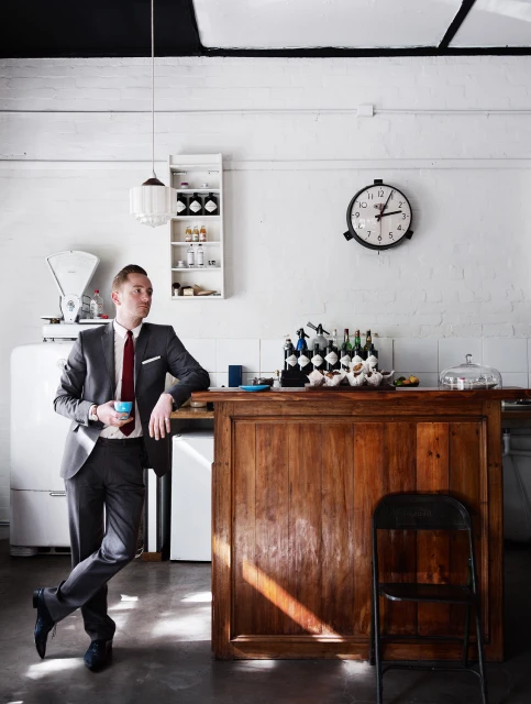 Man in suit leans against wood bar indoors with coffee cup, flanked by vintage scale and bottles, under pendant light and black ceiling.
