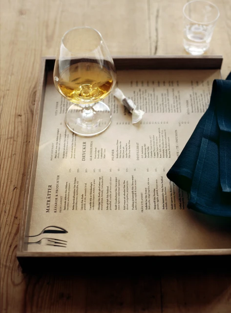 A stem glass of amber liquid sits on a wooden tray with menu and napkin, next to a glass of water on a wooden surface. 