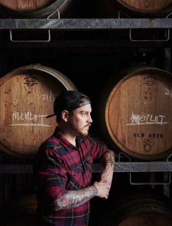 Man with tattoos in a red plaid shirt, standing in a winery, next to barrels labelled 'Merlot' on metal racks in a dimly lit cellar.
