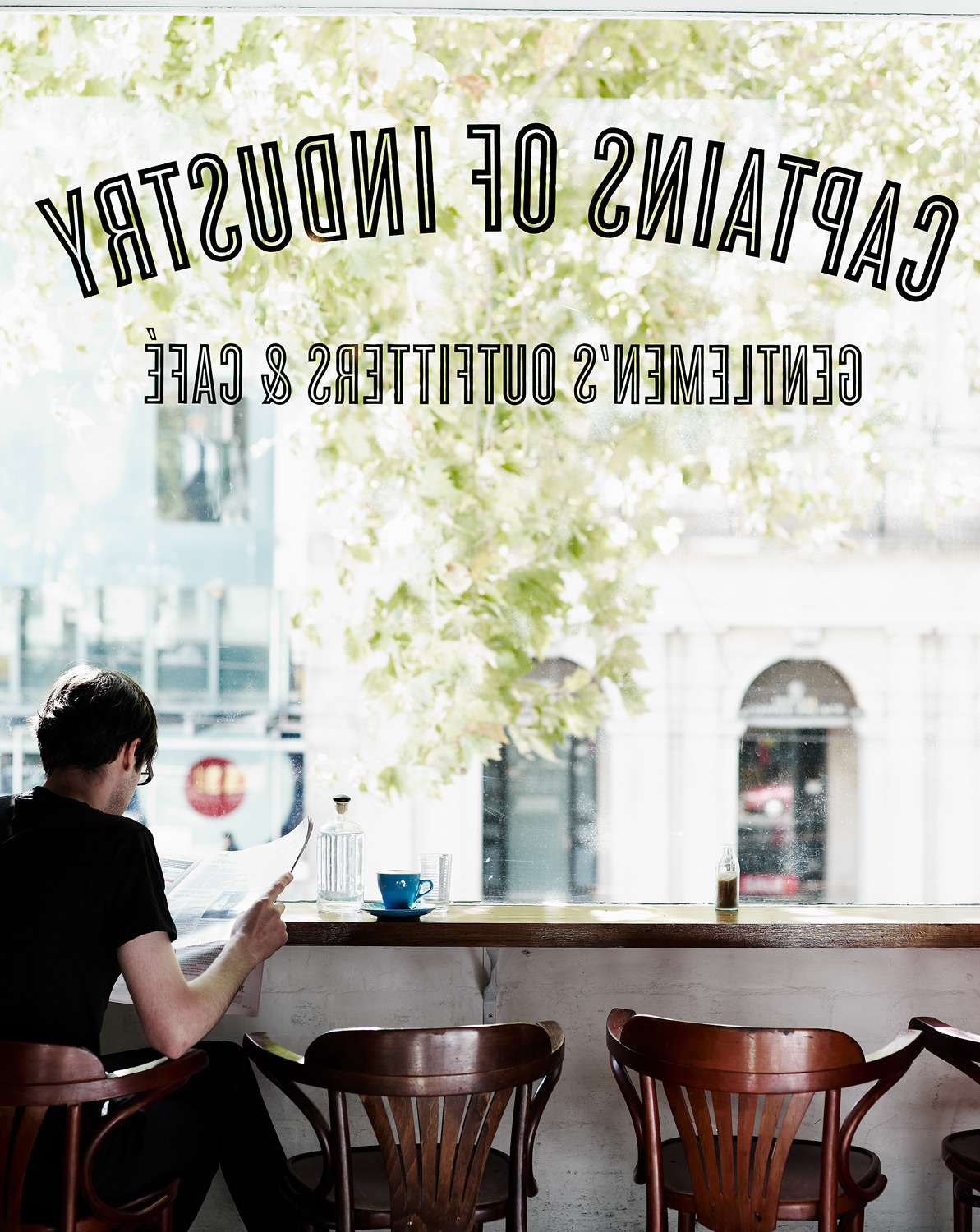 Person reading newspaper at cafe with wooden chairs, tables, and window view of city and greenery. Coffee cup on counter.