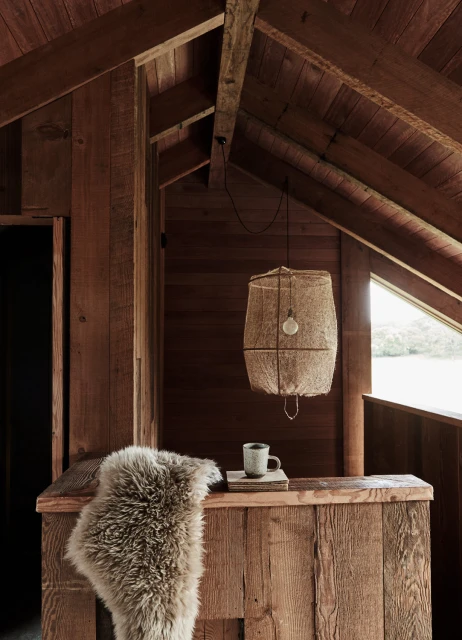 Rustic interior of a wooden cabin featuring a hanging lamp, a fur throw, a small table setting, and a view of a lake through the window.