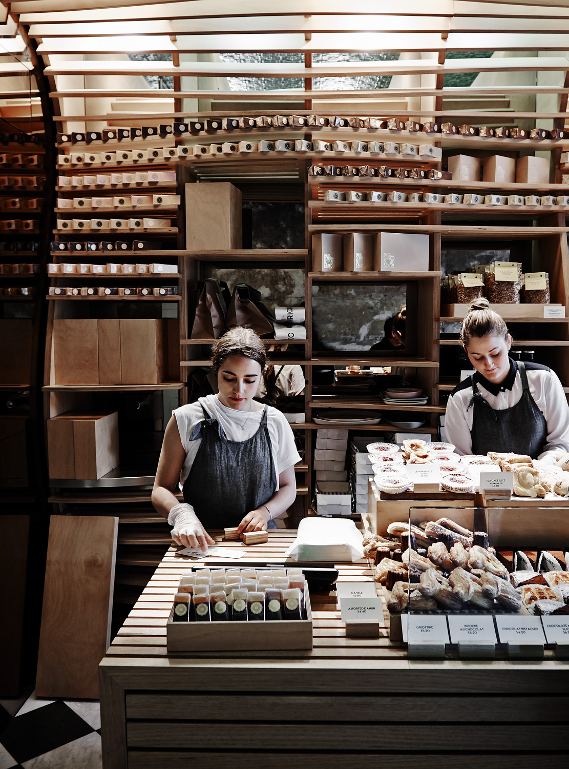 Two women in aprons stand behind a wooden counter with bakery goods and merchandise displayed in a shop with wooden shelves and lighting.