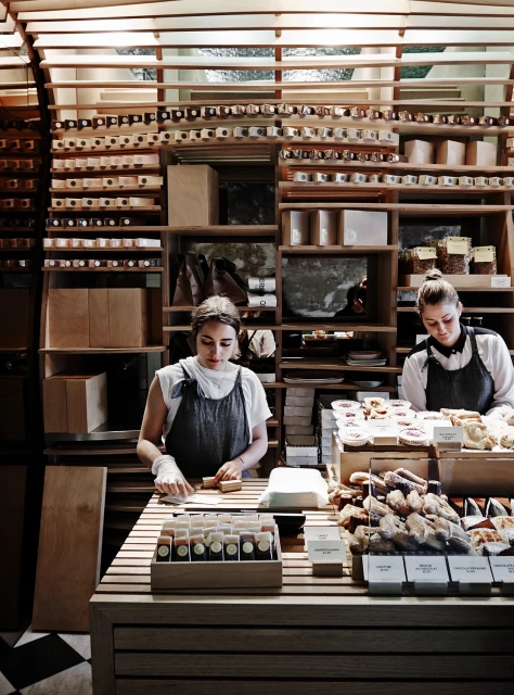 Two women in aprons stand behind a wooden counter with bakery goods and merchandise displayed in a shop with wooden shelves and lighting.