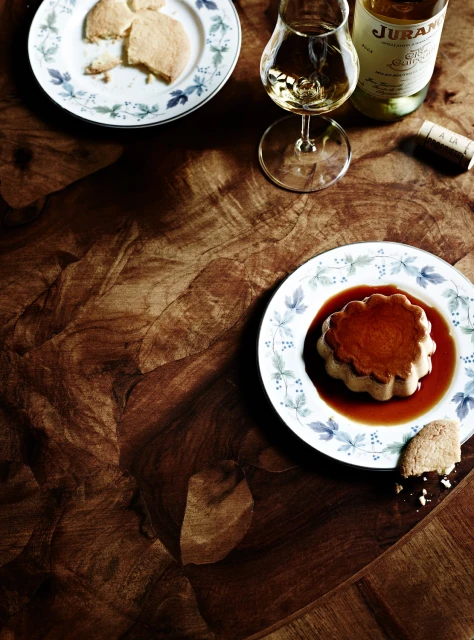 Overhead shot of flan dessert on a plate, accompanied by shortbread cookies, a wine glass, and a bottle of wine on a wood table.