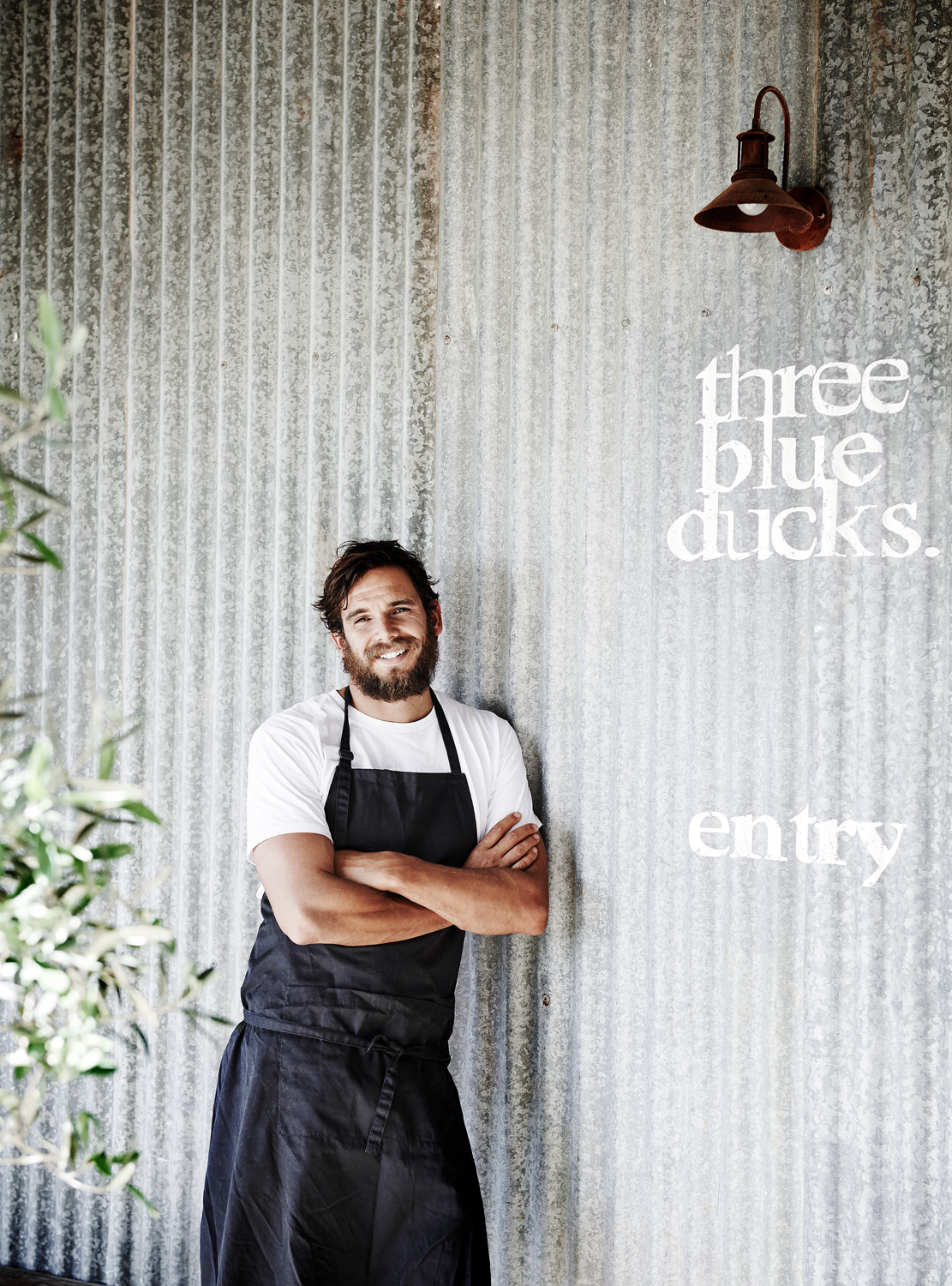 Smiling chef in black apron stands near corrugated metal wall outside with restaurant signage and an outdoor light fixture.