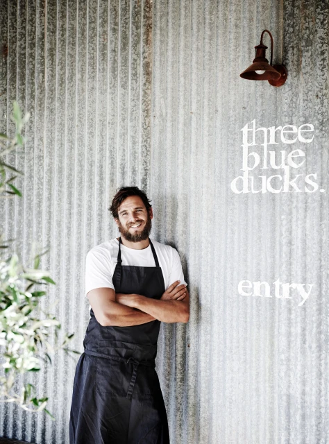 Smiling chef in black apron stands near corrugated metal wall outside with restaurant signage and an outdoor light fixture.