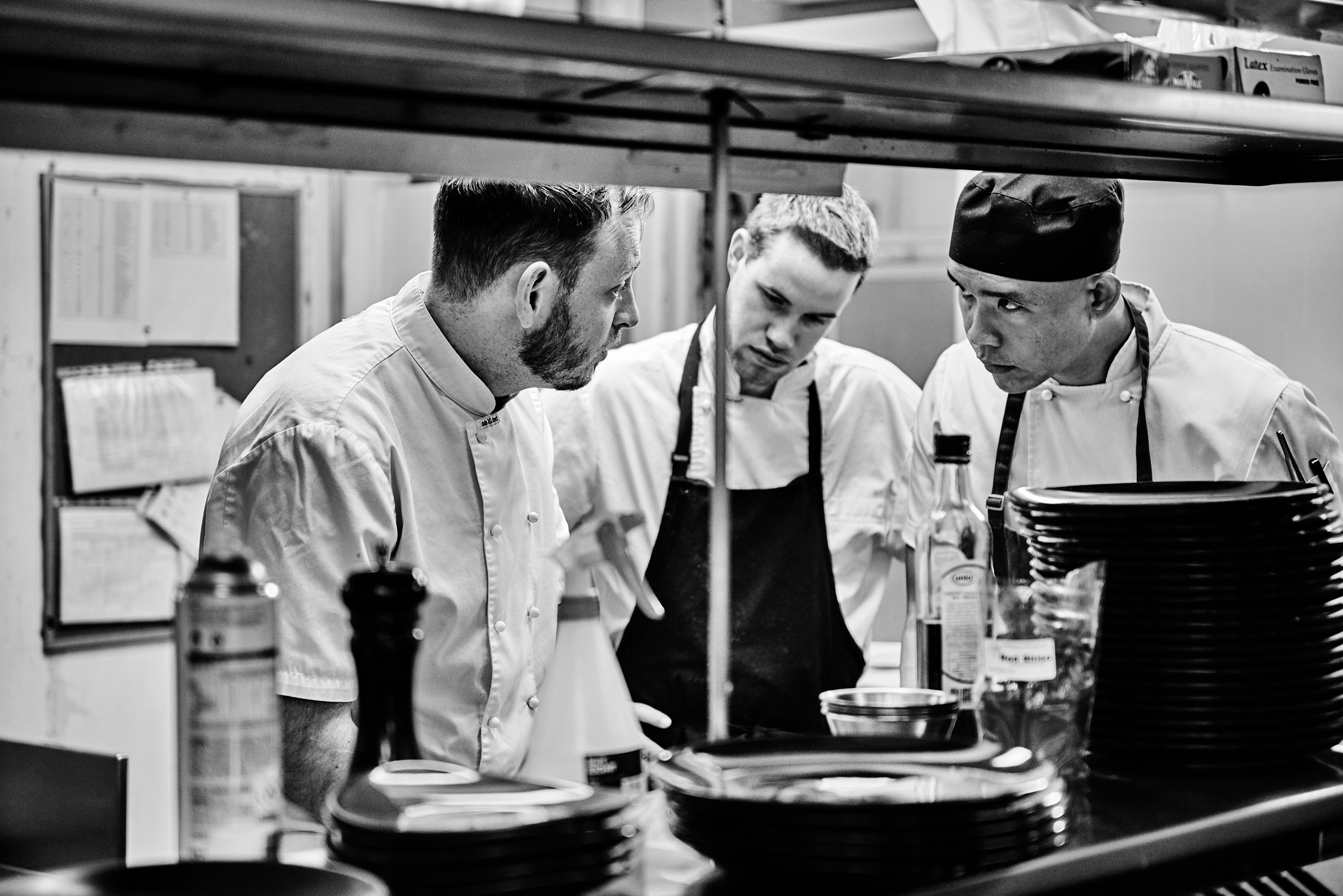 Three chefs in a commercial kitchen are focused on a task. All are wearing chef's whites and aprons in a high contrast black and white image.