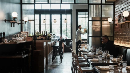 Interior shot of a restaurant with wooden tables, wine bottles on a bar counter, and spherical pendant lights.