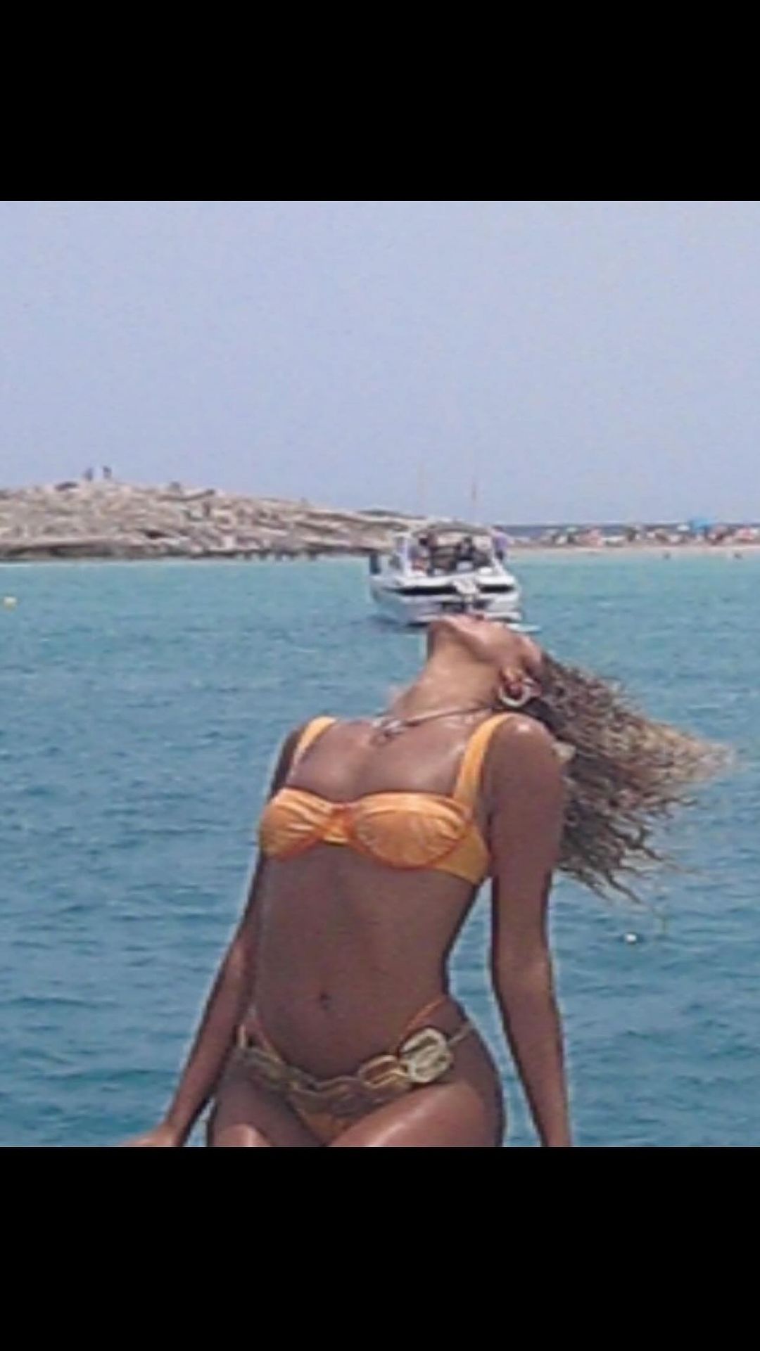 A woman in a yellow bikini poses on a boat against a backdrop of the blue ocean and sky with small islands in the distance.