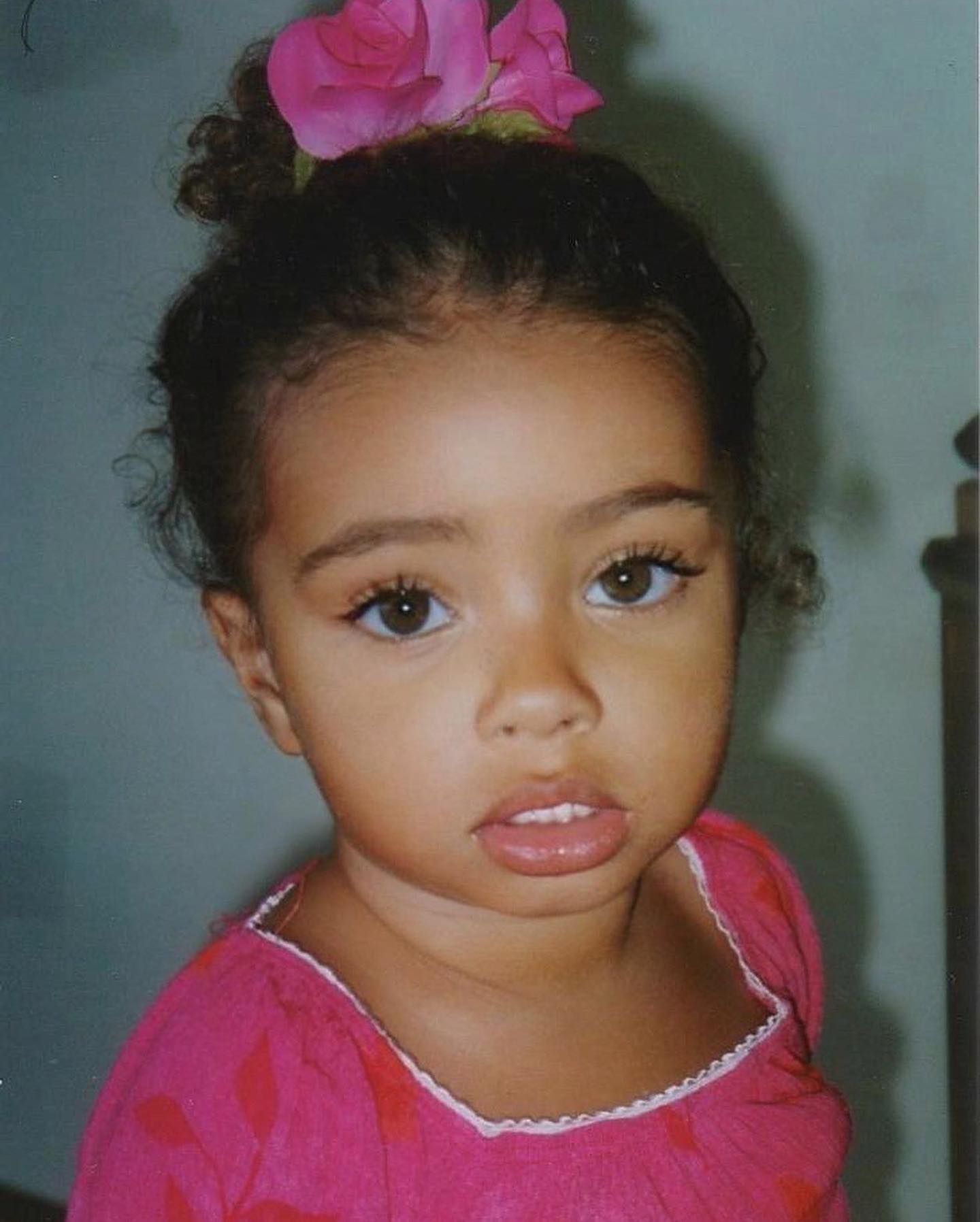 Close-up portrait of a young child with brown eyes and a pink flower in her hair, wearing a bright pink top against a neutral background.