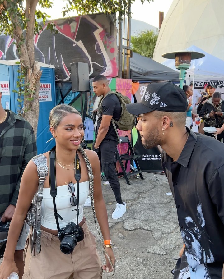 Outdoor shot of a woman with a camera talking to a man in a baseball cap in an urban outdoor setting with other people.