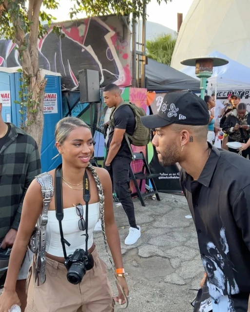 Outdoor shot of a woman with a camera talking to a man in a baseball cap in an urban outdoor setting with other people.