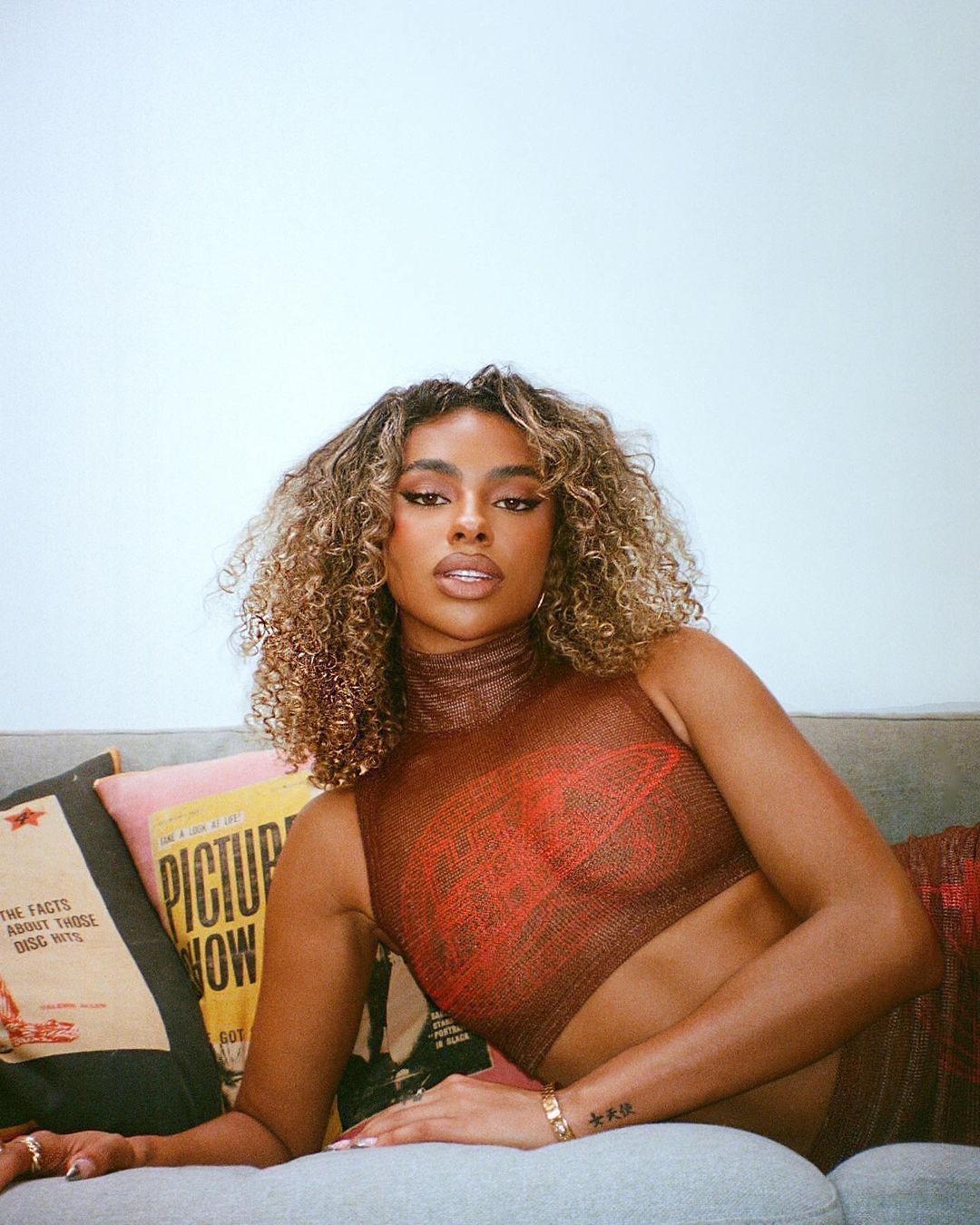 Portrait of a young woman with curly hair posing on a sofa with decorative pillows, wearing a semi-sheer top, indoors.