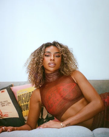 Portrait of a young woman with curly hair posing on a sofa with decorative pillows, wearing a semi-sheer top, indoors.