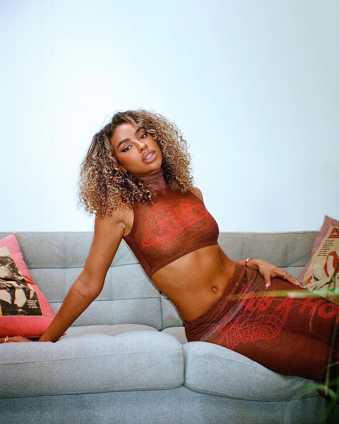 Young woman with curly hair poses on a grey couch in a brown patterned top and pants, illuminated by soft lighting against a white background.