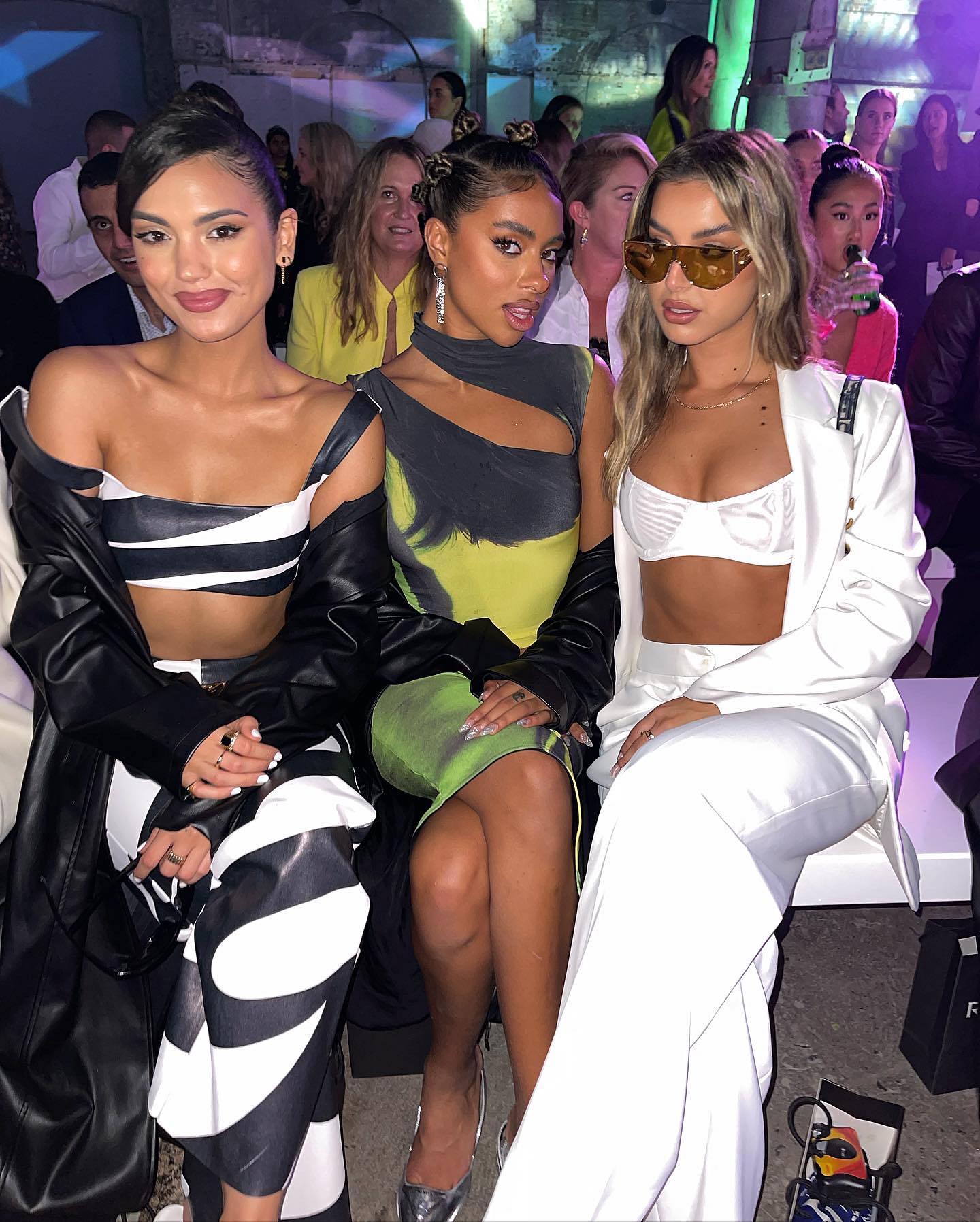 Three women in stylish outfits pose together at an indoor fashion event, showcasing modern designs and runway-ready looks.