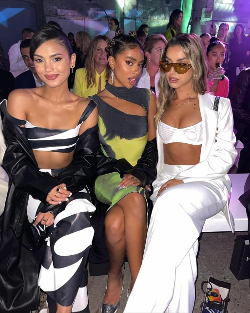 Three women in stylish outfits pose together at an indoor fashion event, showcasing modern designs and runway-ready looks.