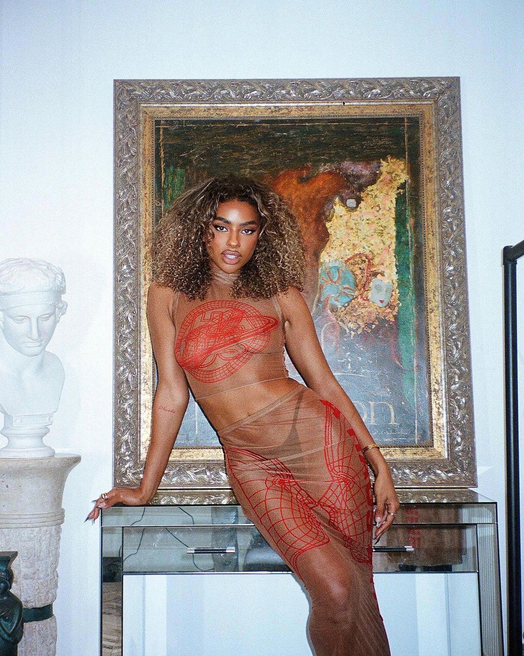 A young adult woman with curly hair poses in a sheer patterned dress, standing near a glass-top table with a marble sculpture backdrop.