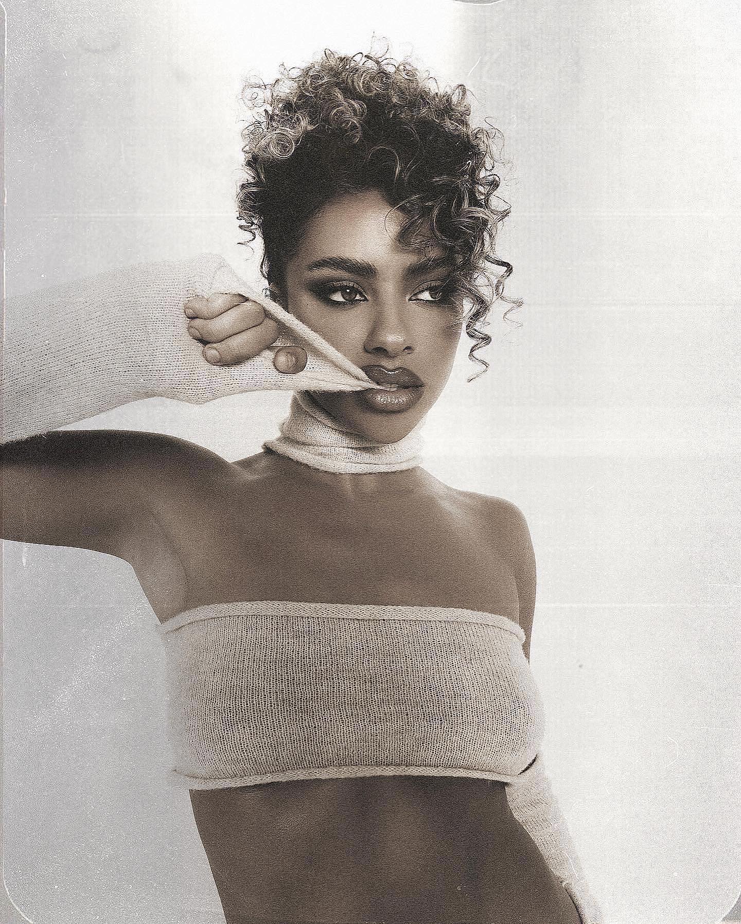 Portrait of an adult female model with curly hair wearing a beige strapless top and scarf, striking a fashionable pose in a photo studio.