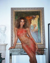 Woman with curly brown hair wearing sheer two piece outfit with red line patterns posing indoors near a framed painting