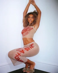 Woman in patterned sheer crop top and matching midi skirt crouching with arms raised against white wall