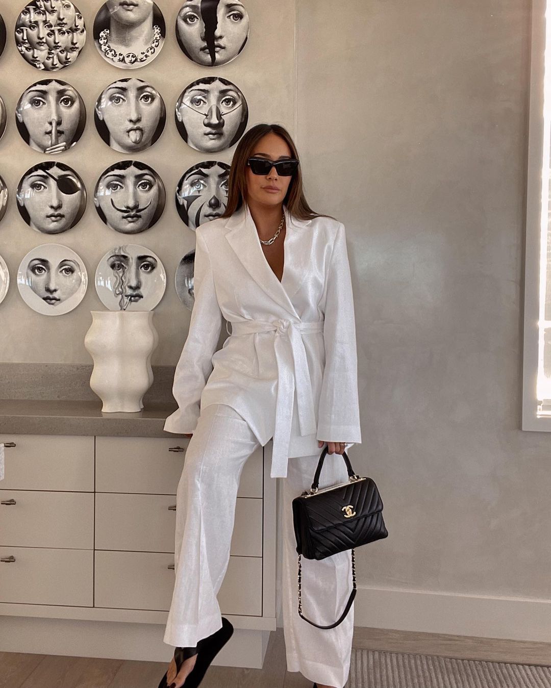 Adult woman poses indoors wearing a white suit and sunglasses while holding a black handbag with gold chain detail.