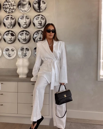 Adult woman poses indoors wearing a white suit and sunglasses while holding a black handbag with gold chain detail.