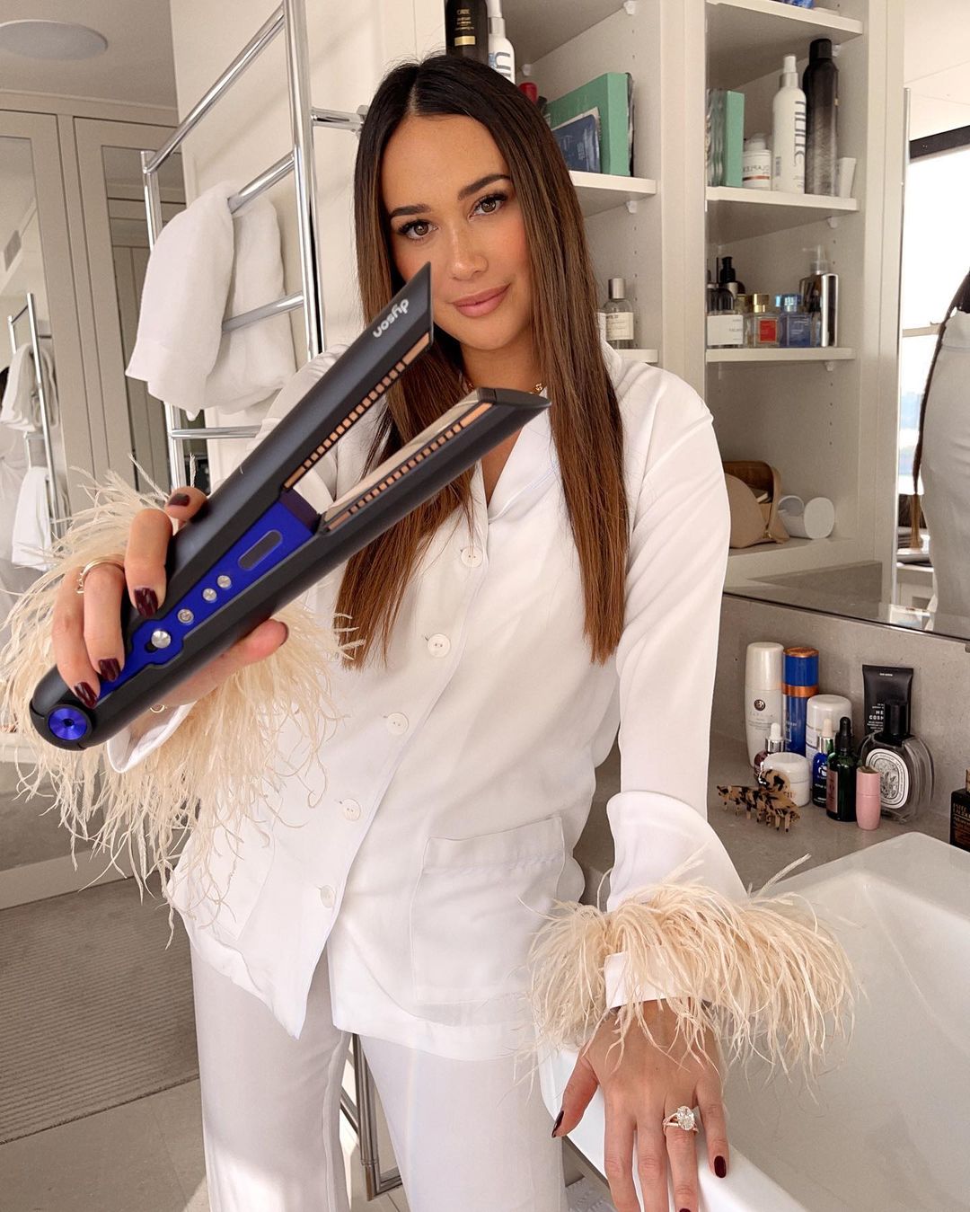 Woman holding Dyson Corrale straightener in bathroom with white pajamas featuring feathered cuffs, showcasing beauty routine.