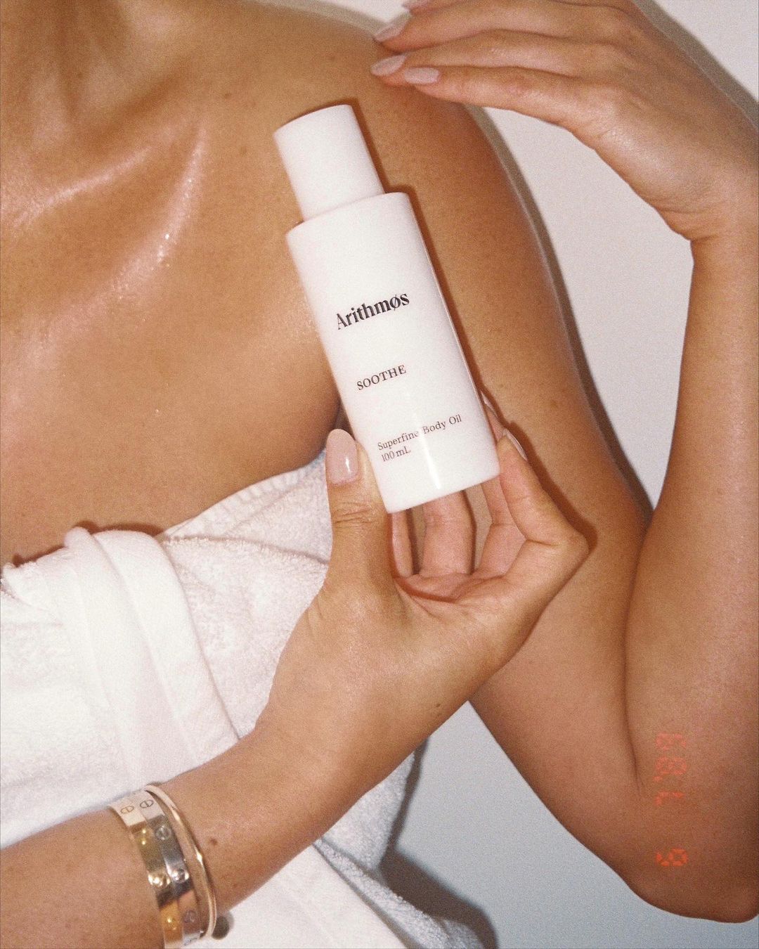 Woman holding a white bottle of Arithmos Soothe Body Oil, partially wrapped in a white towel, against a white background, studio lighting.