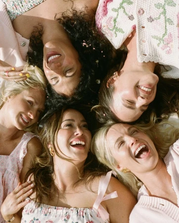 Overhead shot of five smiling women of mixed ethnicities in light-colored summer dresses and tops laying in a circle outdoors.