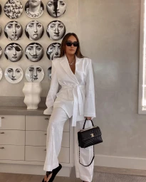 Woman wearing a white suit and black sunglasses holding a quilted handbag against a decorative plate wall