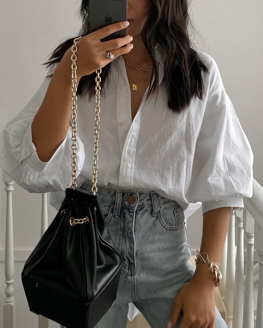 Woman takes mirror selfie wearing a white blouse, jeans, gold jewelry, and holding a black purse with a gold chain handle.