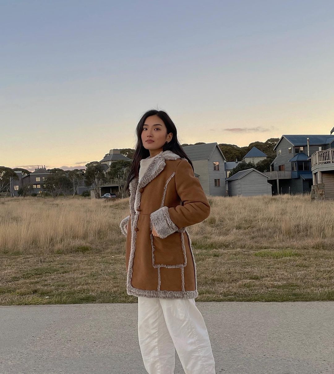 Asian woman poses outdoors wearing brown suede coat with white lining and trim, and loose white pants against a rural landscape.
