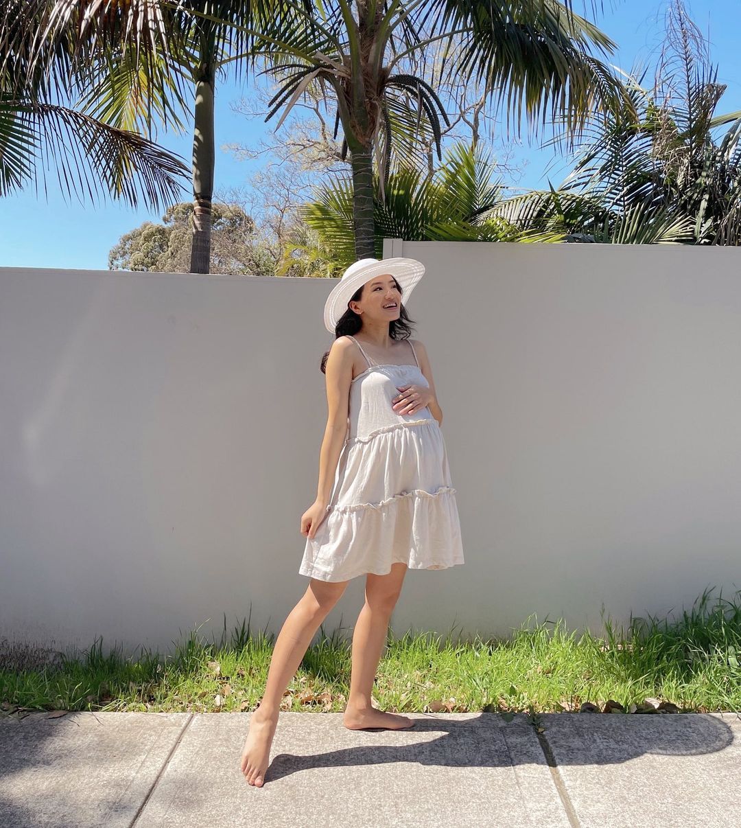 Pregnant woman in a sundress and hat stands barefoot in front of a white wall and palm trees on a sunny day in an outdoor setting.