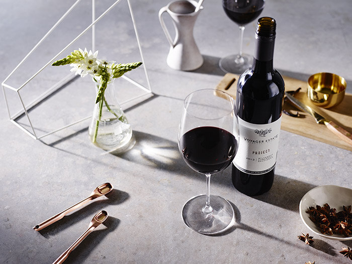 Still life shot of a wine bottle and glass, a vase of flowers, copper spoons, and a gold cup on a gray surface.