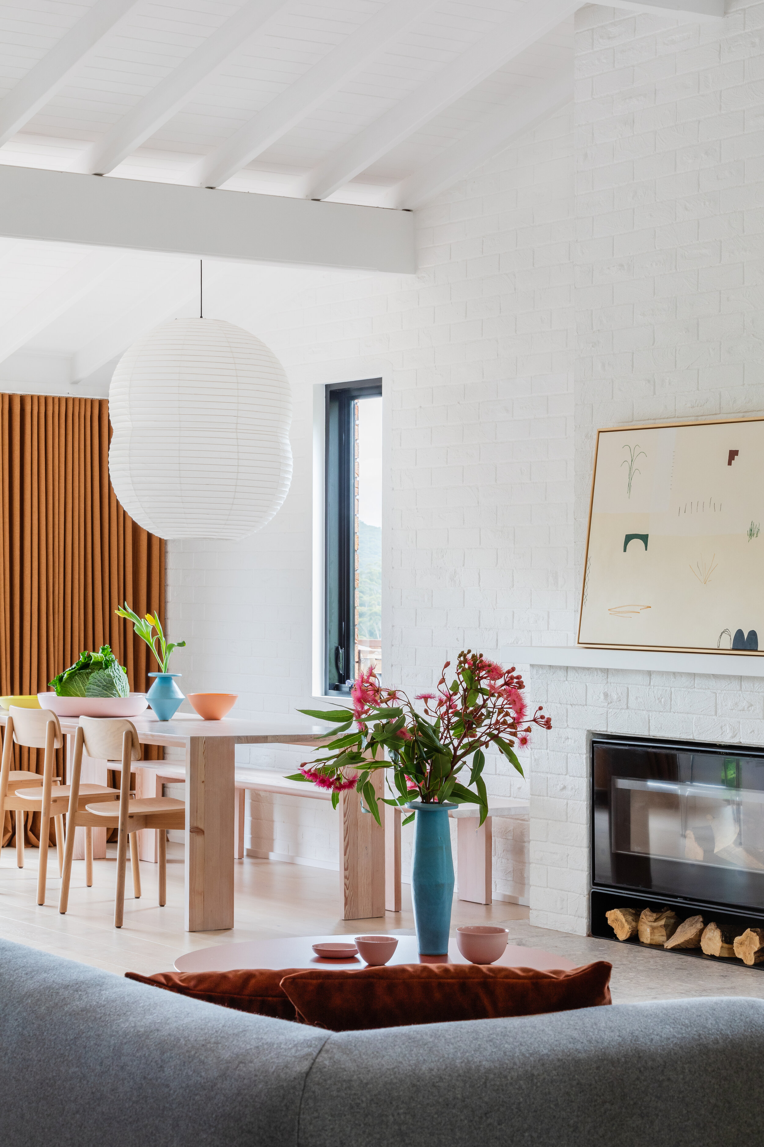 Living room interior with white brick walls, dining table with chairs, modern fireplace, grey sofa, and vase of red flowers.