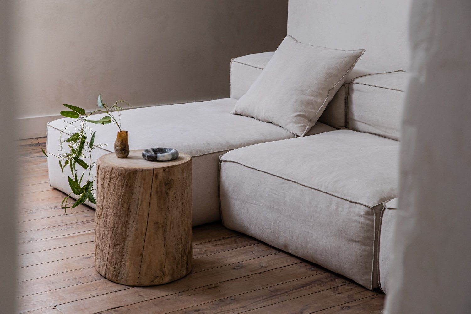 Living room corner features a modular sofa, natural wood stump table with a vase, ring, and botanical sprig.