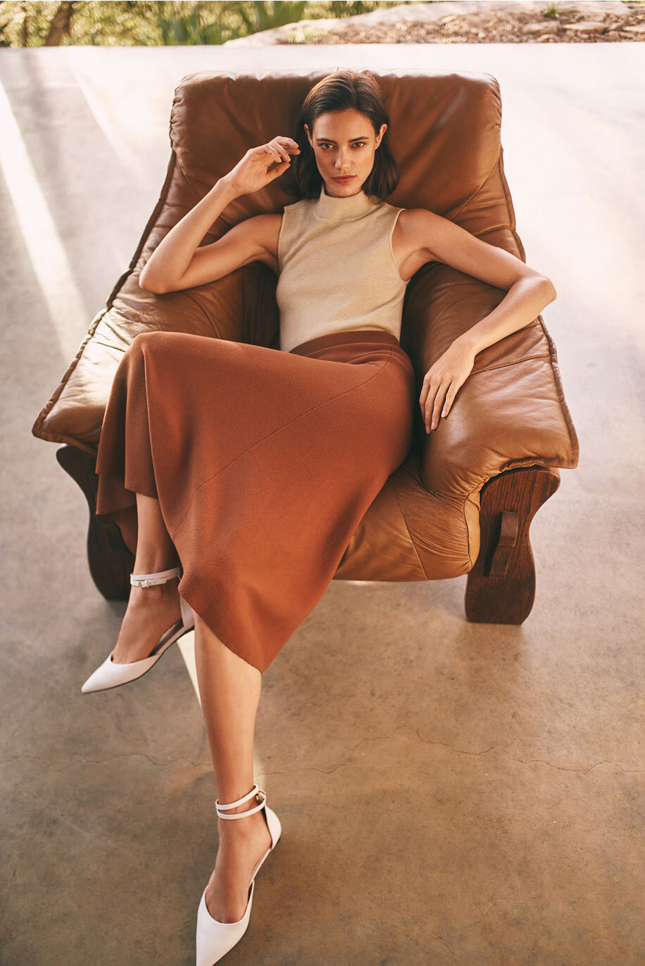 A woman sits in a brown leather chair wearing a beige top, brown skirt, and white high heels in an indoor setting with natural light.