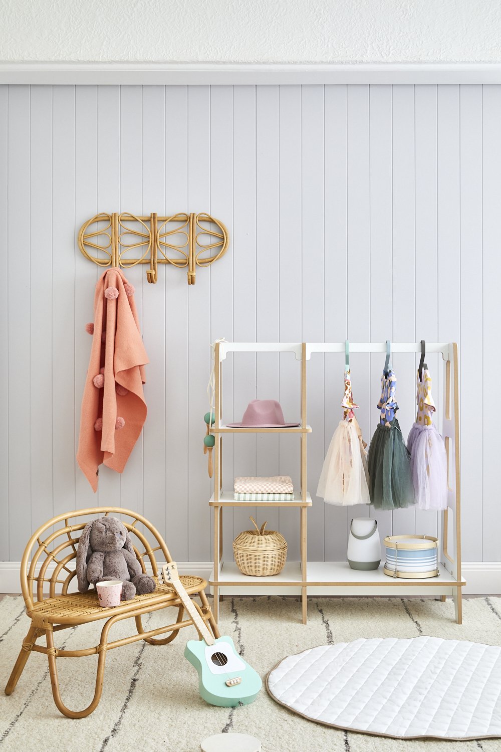 Rattan furniture and playful decor in a children's room with a wooden clothing rack, toy guitar, stuffed animal, and soft neutral colors.