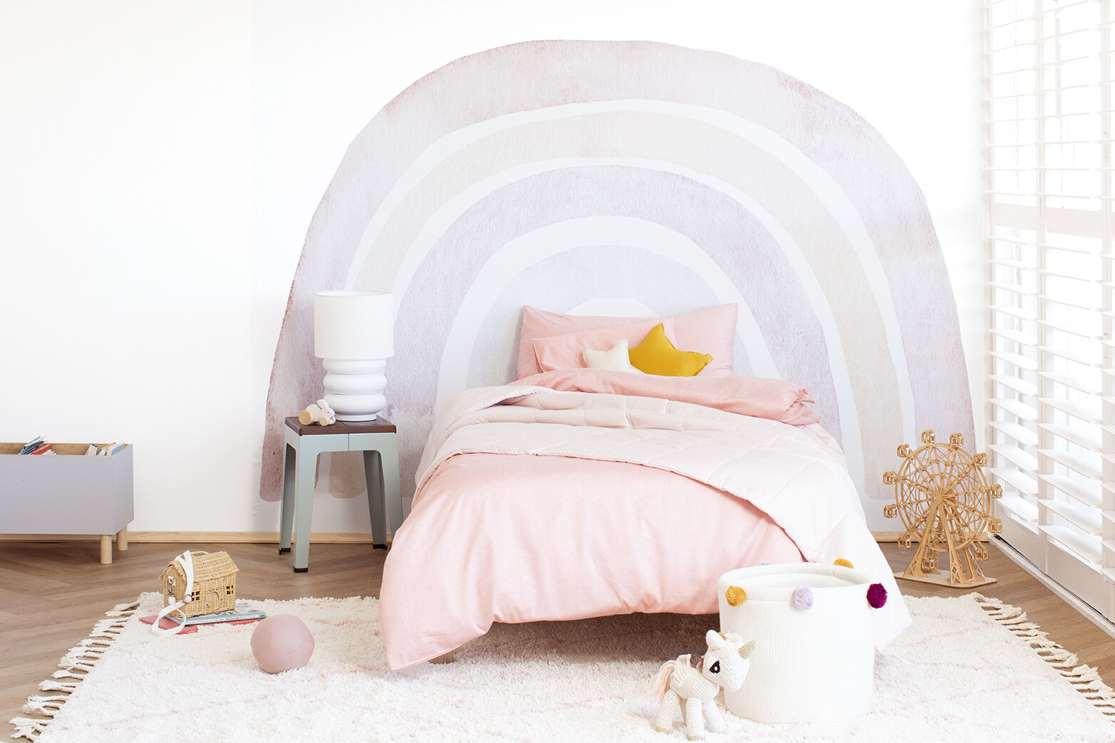 Cozy kids' bedroom with pink and white bedding, rainbow wall decoration, and various toys, creating a playful and inviting space.