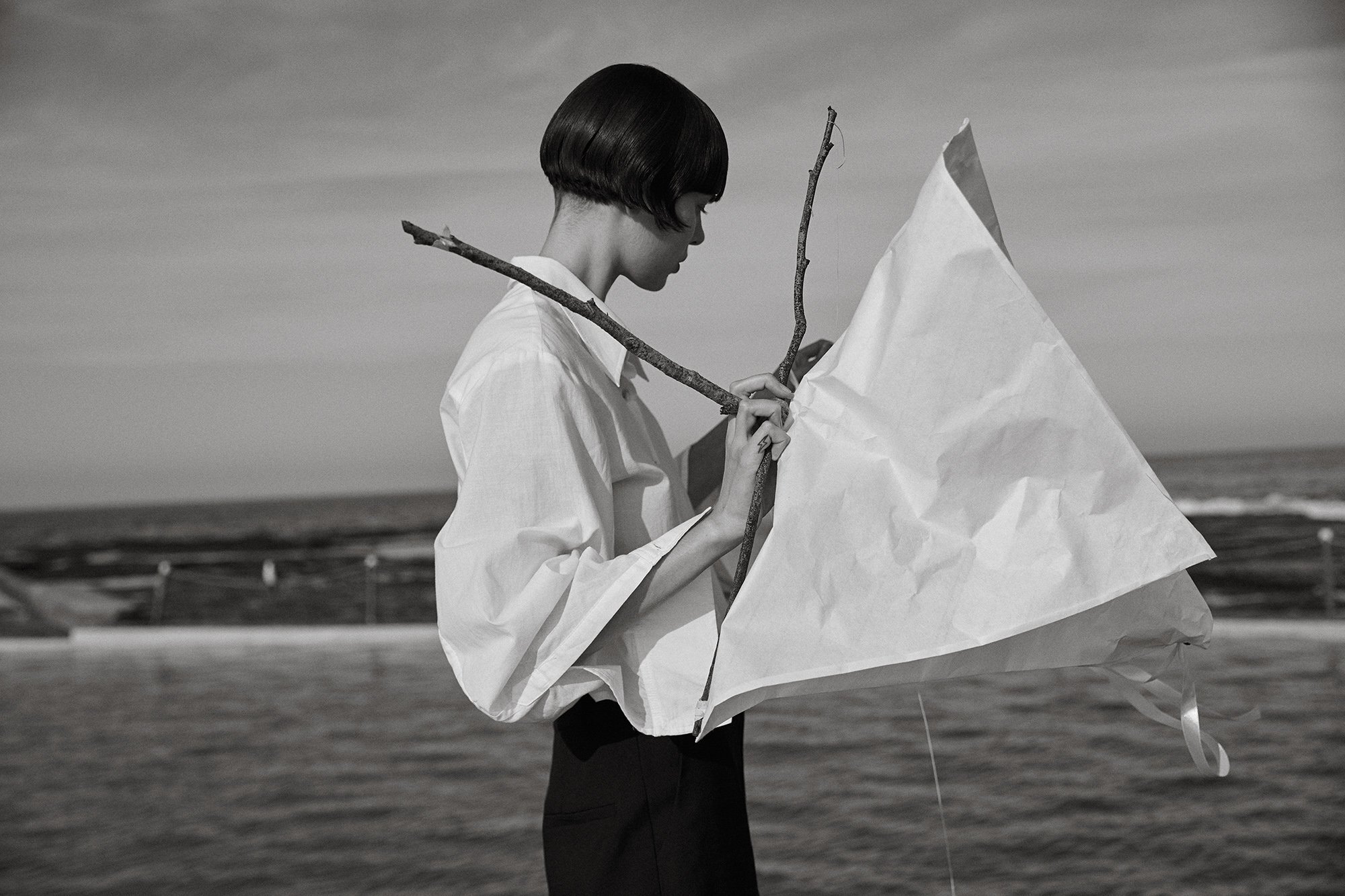 Black and white shot of woman with short bob holding sticks and a white cloth on a beach with ocean and sky background.