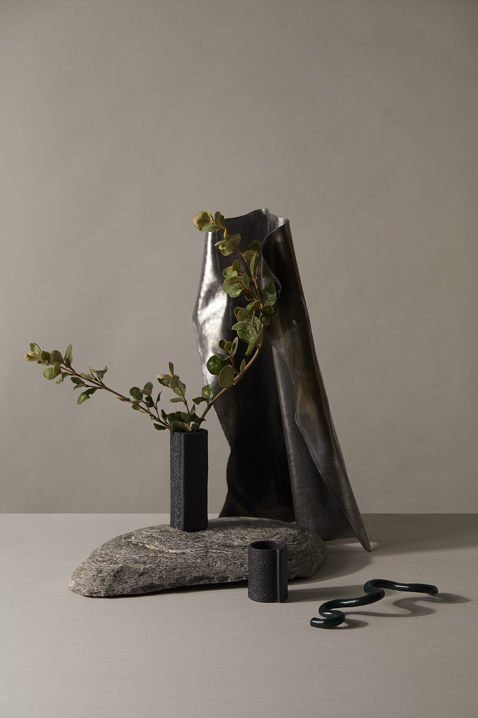Still life composition featuring black vases, green plant sprigs, a grey rock, and a silver fabric backdrop against a neutral background.