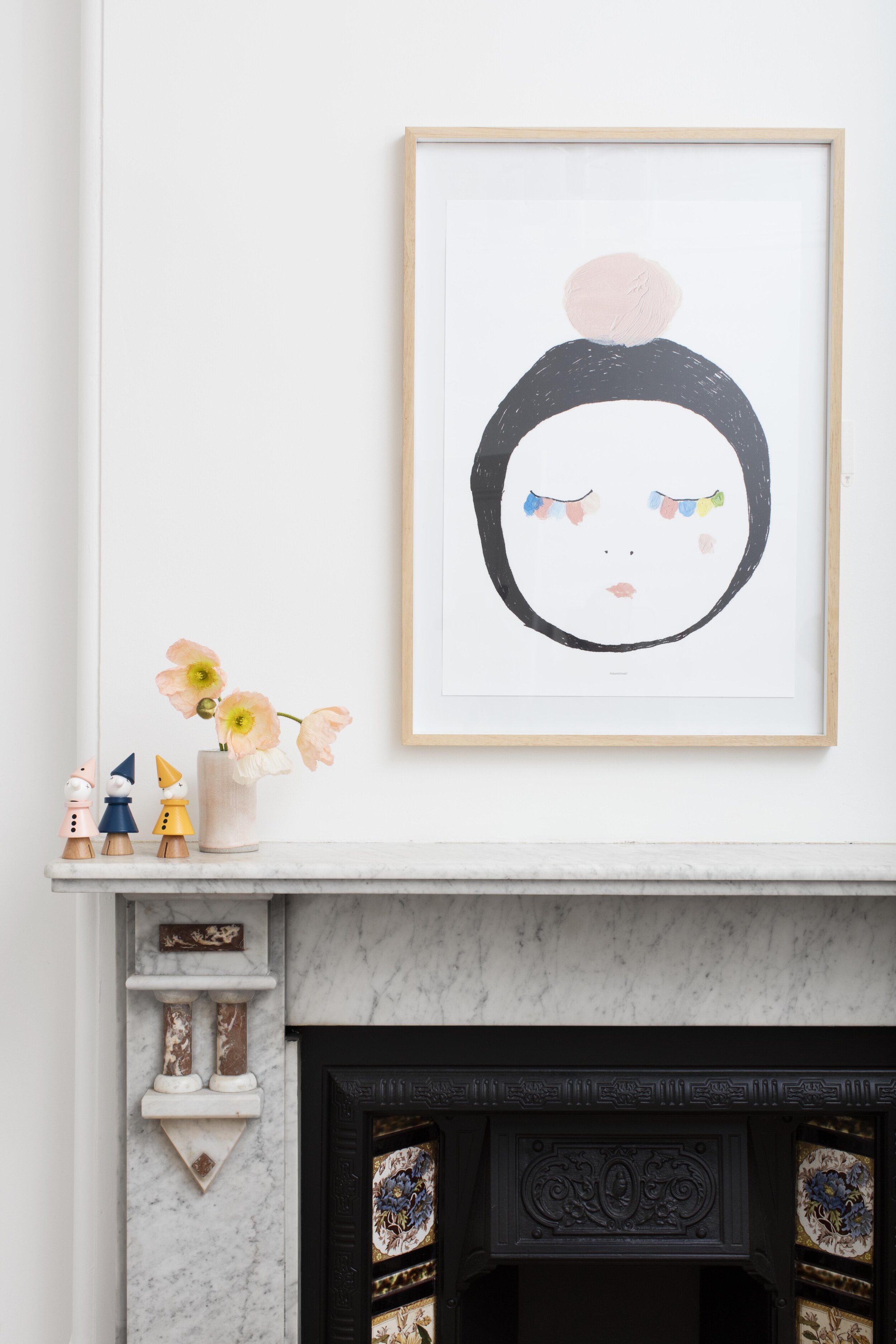 Framed illustration of a face on a marble fireplace mantel, adorned with wooden toys and a vase of peach-colored flowers.