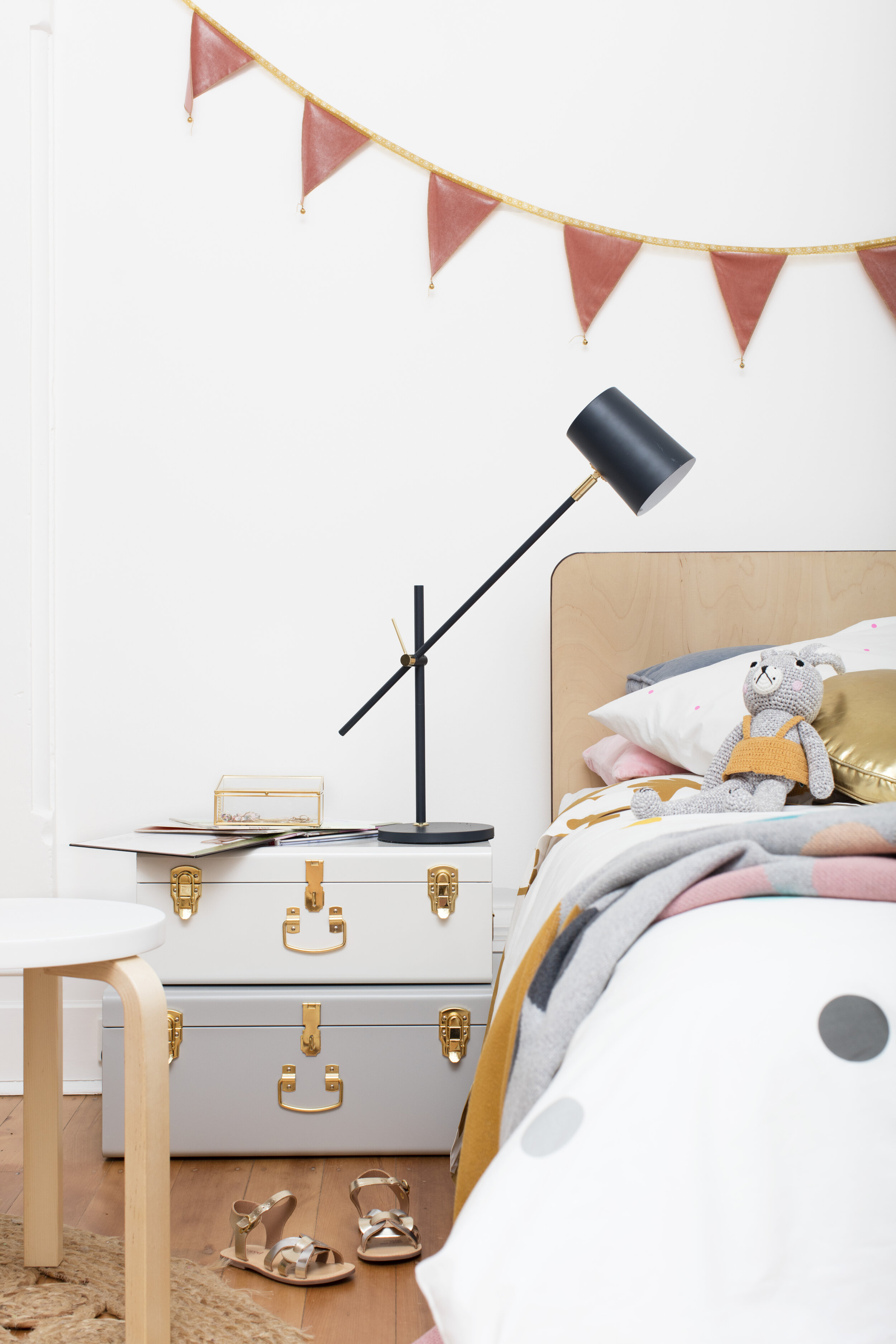 Interior of a child's bedroom with a bed, nightstand made of stacked storage trunks, table lamp, and string of pennant flags.
